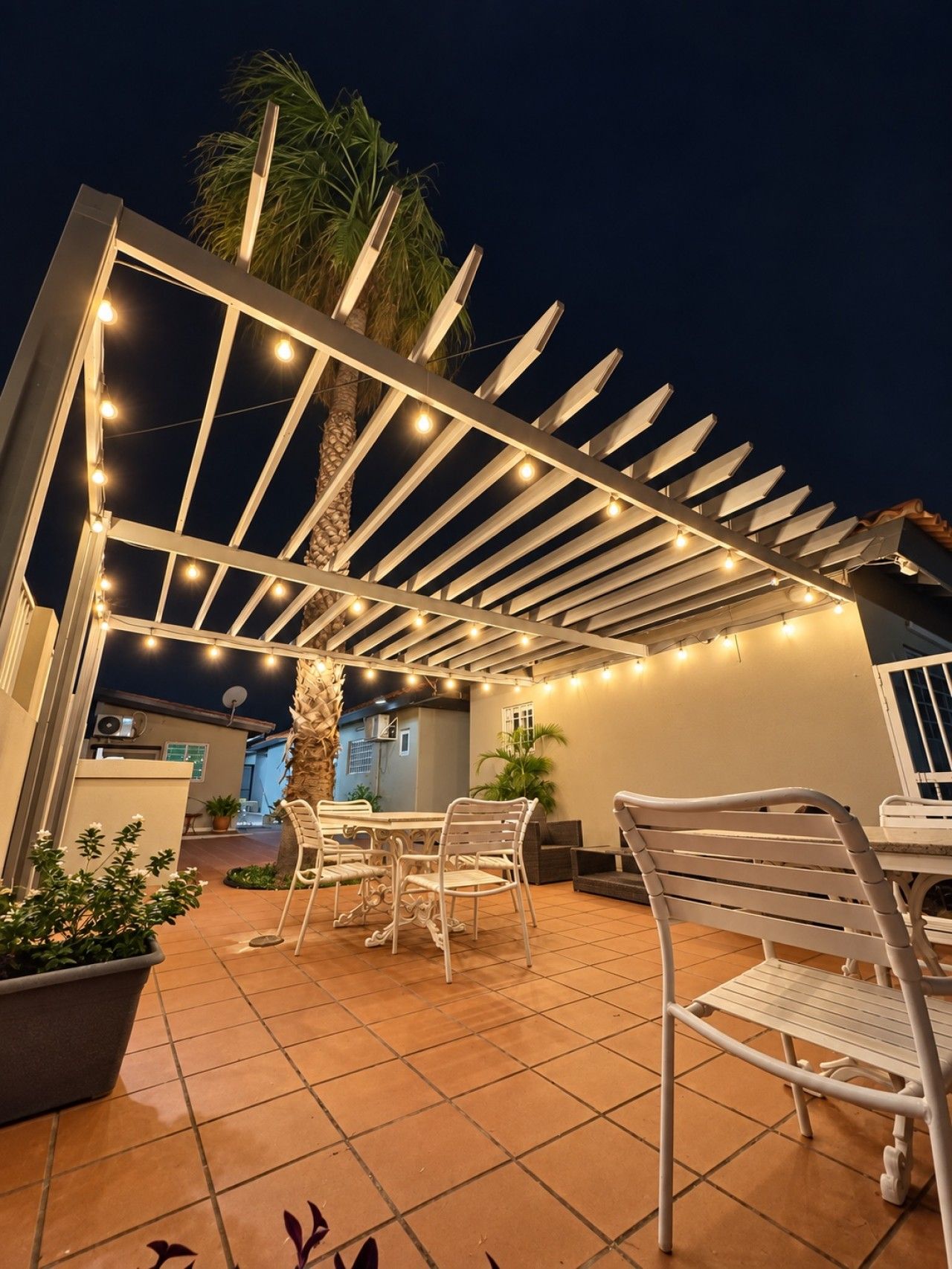 Een terras buiten met een pergola, lichtslingers en tuinmeubilair.