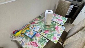Keittiössä on kaasuliesi, pöytä, jonka päällä on kukkakuviollinen pöytäliina, sekä kaksi tuolia.