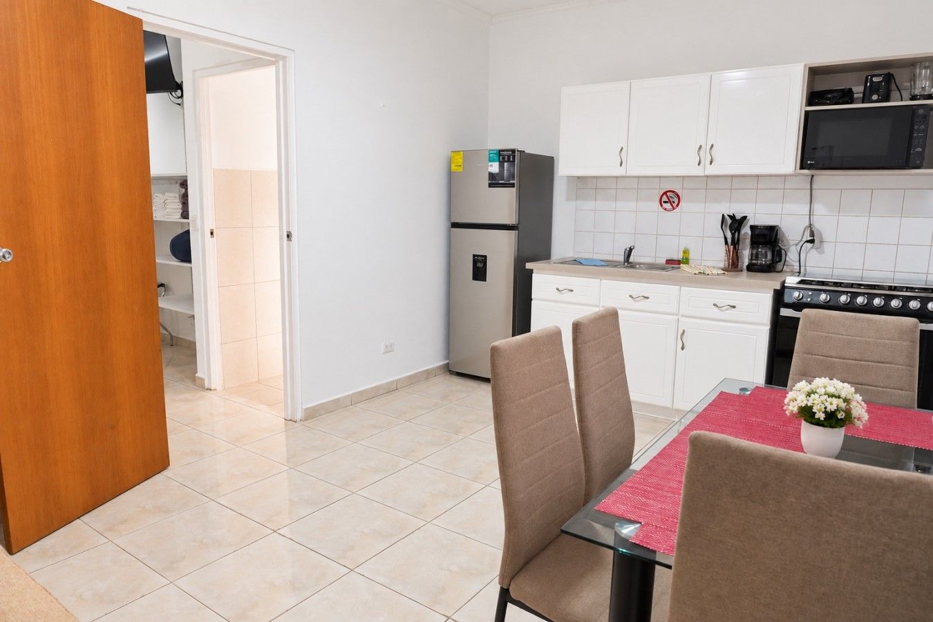 Een kleine keuken met witte kasten, een koelkast, een fornuis en een eettafel met stoelen.