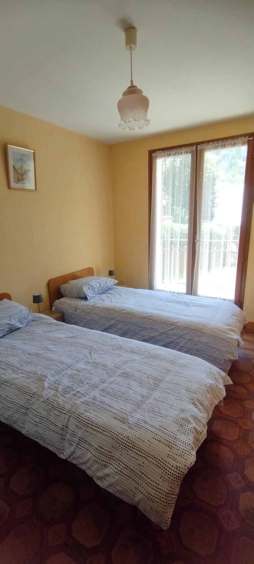 A room with two beds, a ceiling light, a window with curtains, and a framed picture on the wall.