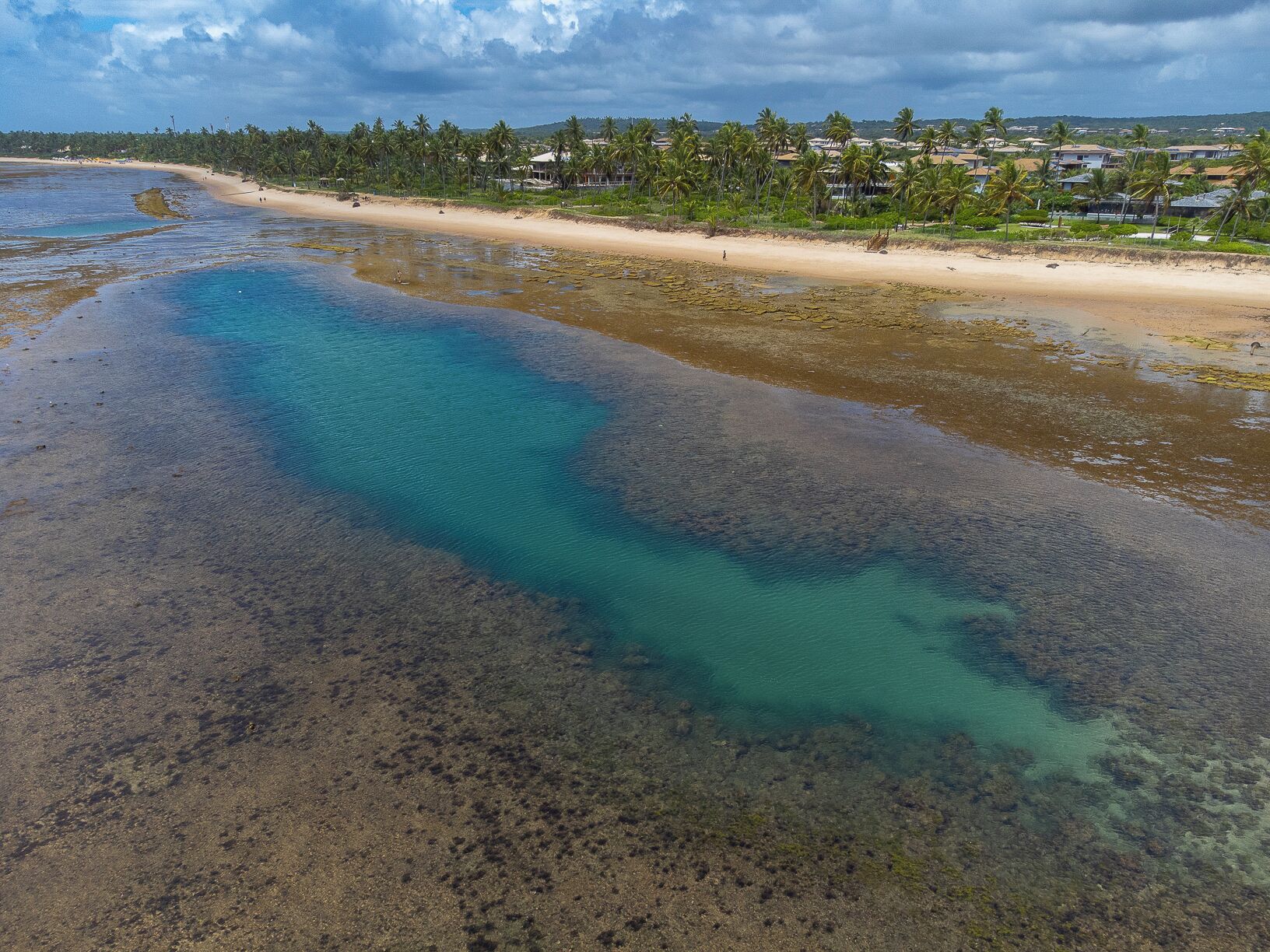 A coastal landscape with a clear water body, sandy beach, and a line of houses in the background.