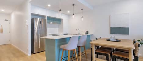 Una cocina moderna con una isla central, electrodomésticos de acero inoxidable, iluminación colgante y un comedor con sillas y mesa de madera.