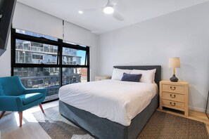 Ein modernes Schlafzimmer mit einem großen Bett, einem blauen Sessel, einem Nachttisch mit Lampe und Blick auf die Stadt durch große Fenster.