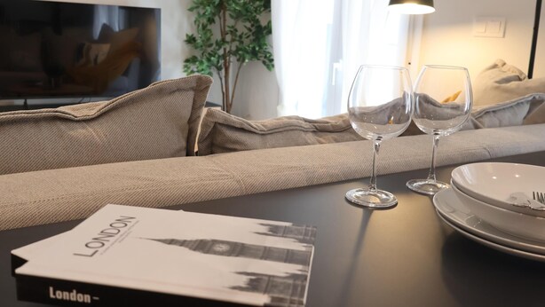 Uma sala de estar moderna com um sofá, dois copos de vinho, um livro intitulado 'LONDON' e um abajur.