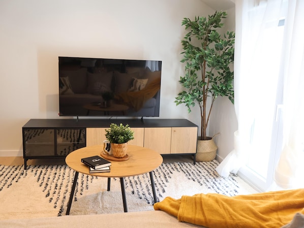A living room with a TV, a wooden cabinet, a round table with books, a potted plant, and a yellow blanket.