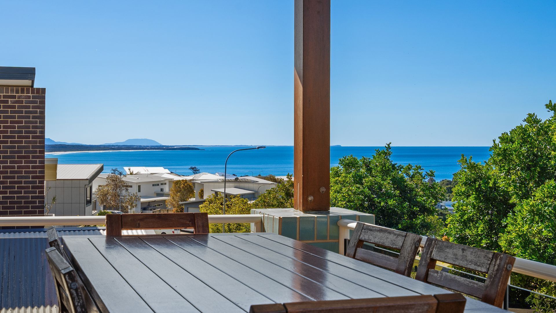 Terraza de madera con mesa y sillas con vistas al mar, edificios y cielo despejado.