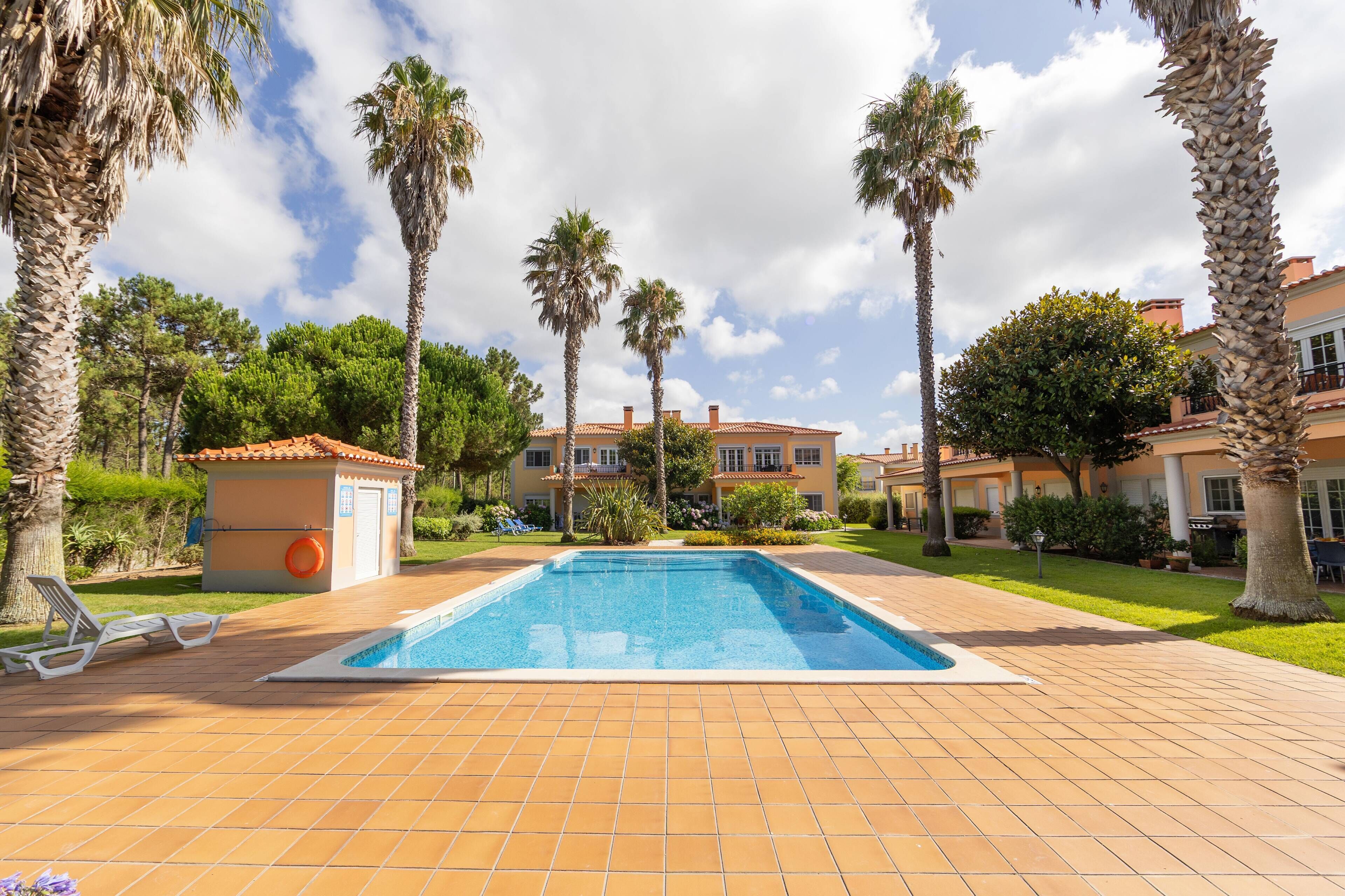 A swimming pool with a tiled deck, surrounded by palm trees and a building with a tiled roof.