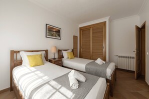 A hotel room with two beds, a nightstand, a lamp, a framed picture on the wall, and a wooden wardrobe. - Destino Praia D'El Rei - Shared Pool (Óbidos)