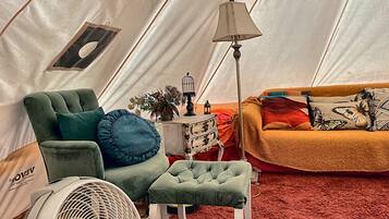 A tent interior with a green armchair, a small ottoman, a white fan, a lamp, and a sofa with patterned cushions.