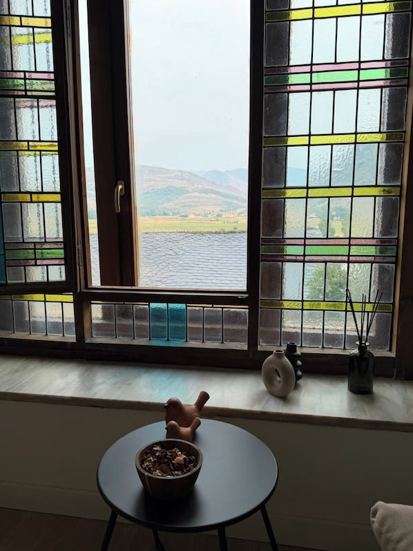 Una habitación con vista a un lago y colinas, una ventana con vistas, una mesita con un cuenco y un florero con ramas.