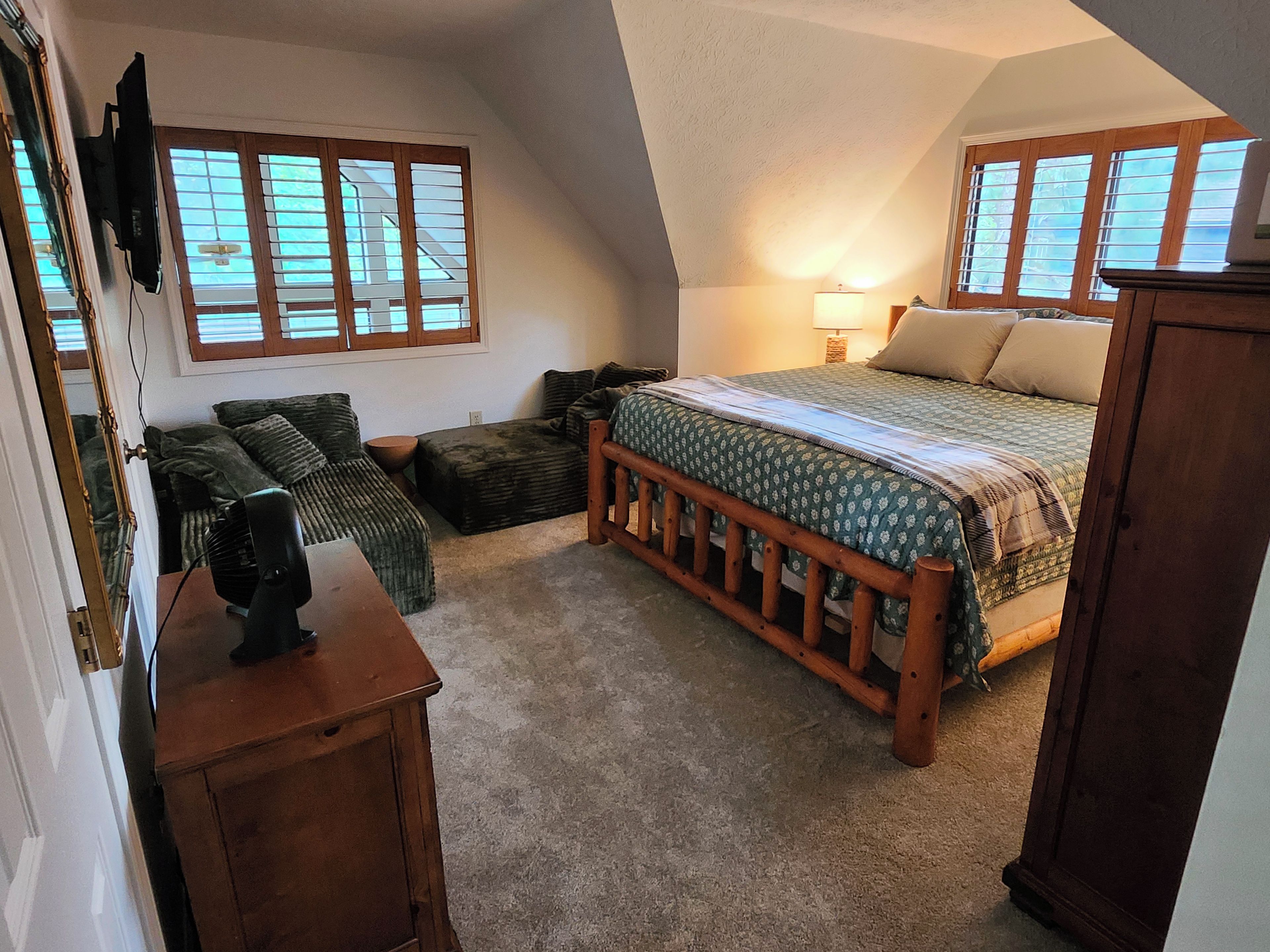 A bedroom with a sloped ceiling, a bed with a wooden headboard, a nightstand, a lamp, and a window with blinds.