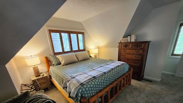 A bedroom with a sloped ceiling, a bed with a patterned bedspread, a wooden dresser, and a window with blinds.