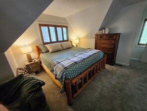 A bedroom with a sloped ceiling, a bed with a patterned bedspread, a wooden dresser, and a window with blinds.