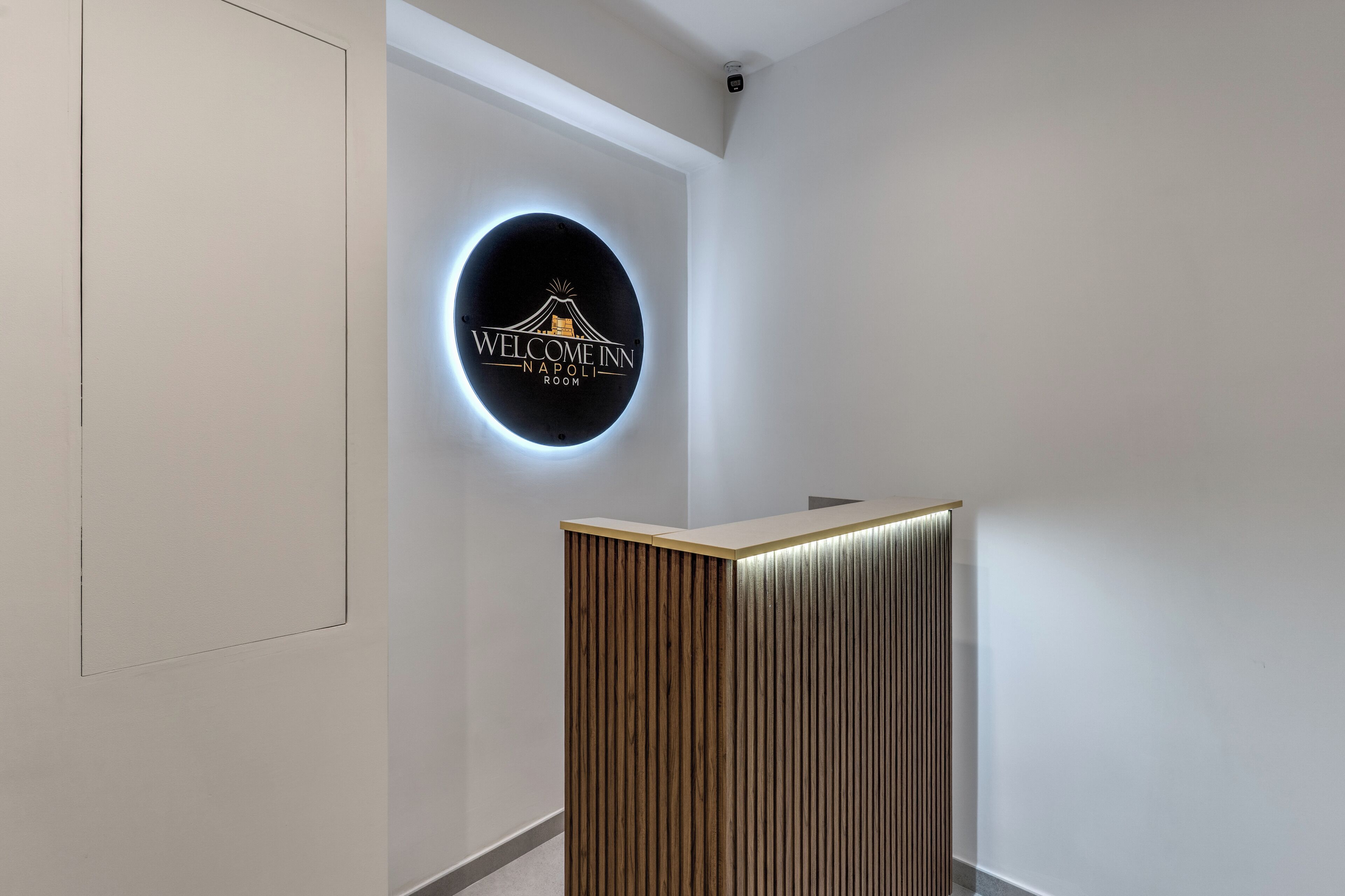 Un'area reception con un cartello circolare che indica 'Welcome Inn Napoli Room' e un bancone reception in legno.