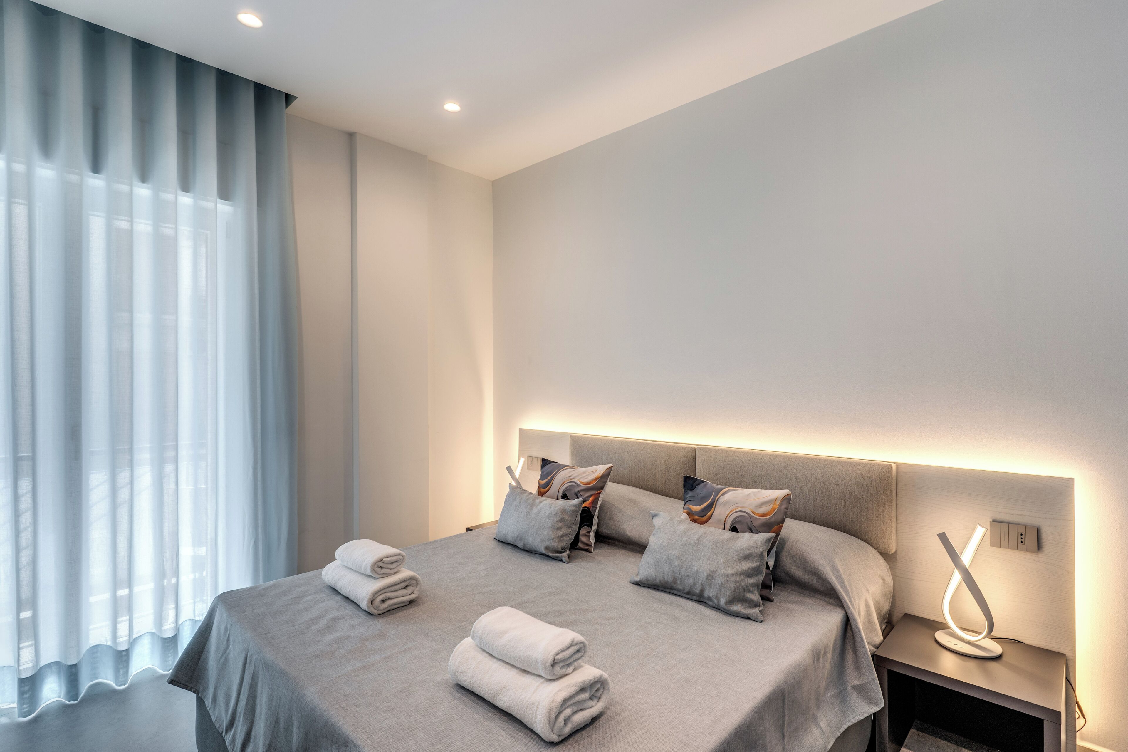 Ein modernes Hotelzimmer mit einem ordentlich bezogenen Bett, einem Nachttisch mit Lampe und einem großen Fenster mit leichtem Vorhang.