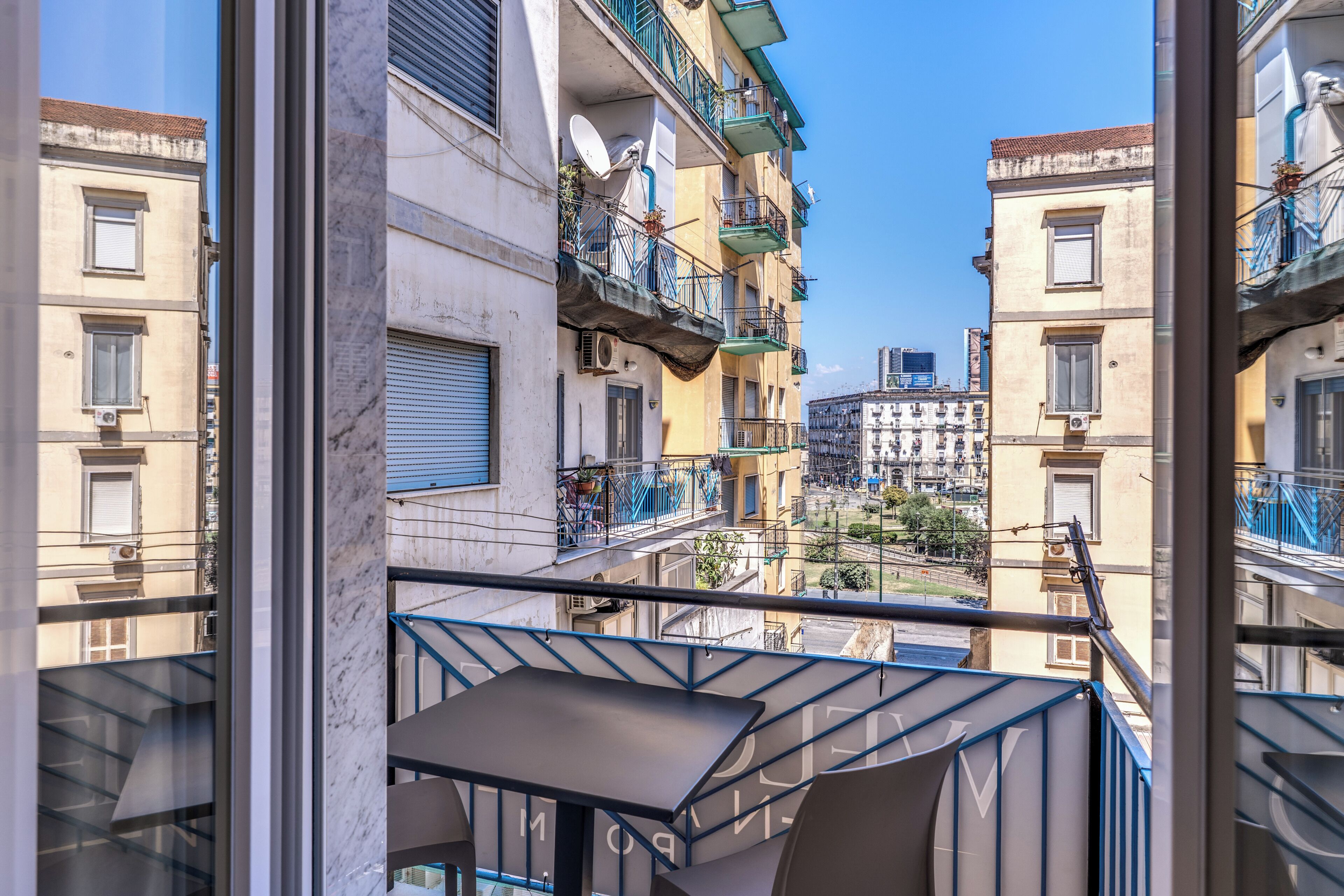 A view from a balcony overlooking a street with multi-story buildings, some with balconies and air conditioning units.