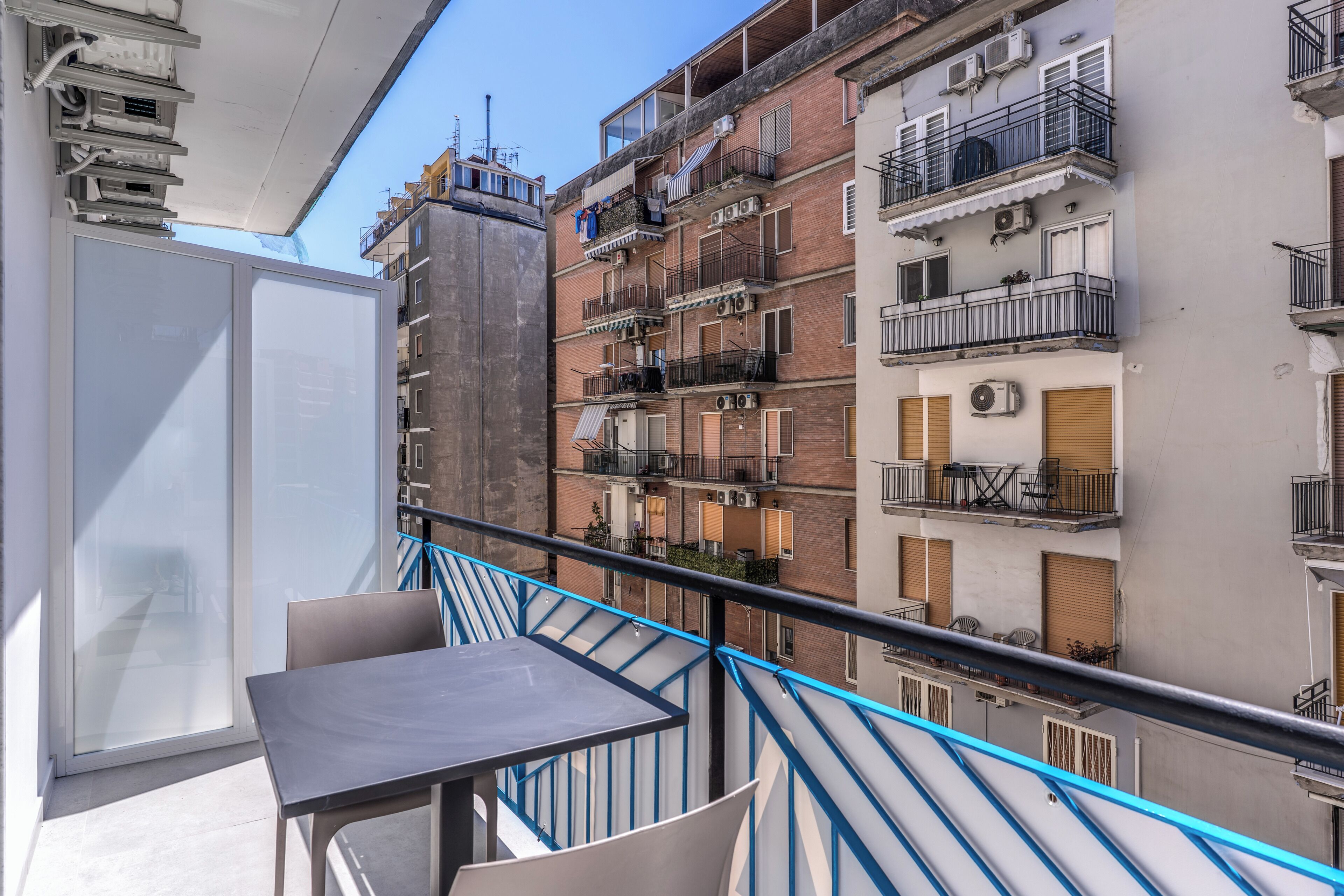 Ein Balkon mit Tisch und Stühlen, mit Blick auf eine Reihe von mehrstöckigen Gebäuden mit Balkonen und Klimaanlagen.