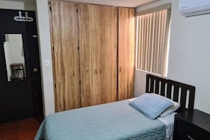 A bedroom with a bed, a wooden wardrobe, a window with blinds, and a small table.