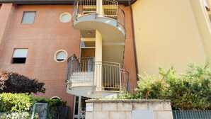 Un immeuble résidentiel à plusieurs étages, doté d’une façade en briques, d’un balcon avec des balustrades en métal et d’un petit jardin agrémenté d’arbustes et d’une clôture.