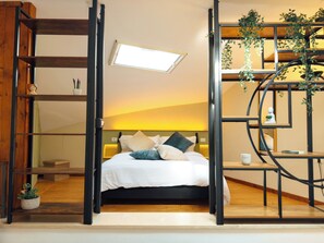 A bedroom with a bed, a bedside table, a nightstand, a window, and a plant.