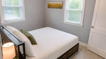 A bedroom with a bed, two windows, a ceiling fan, a bedside table with a lamp, and a door.