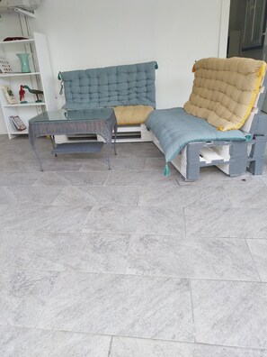 A room with a grey tiled floor, a glass-top table, a blue and yellow sofa, and a white shelf with decorative items.
