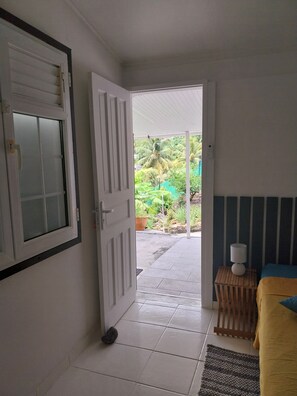 A room with a bed, a bedside table with a lamp, a window with blinds, and a door leading to an outdoor area with a view of greenery.