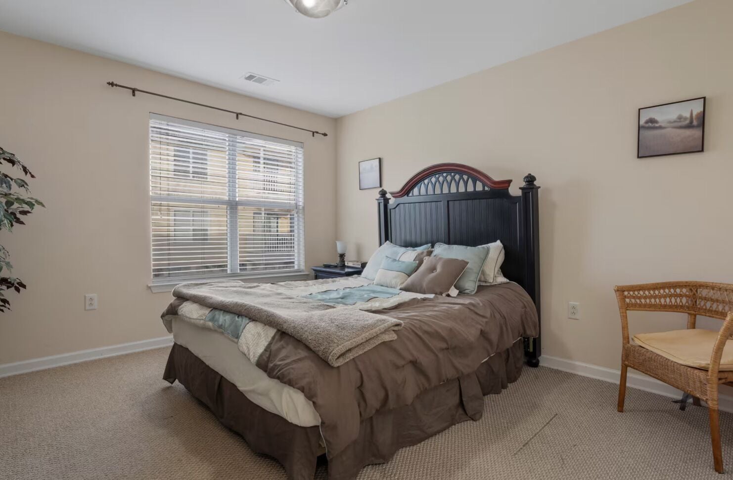 A bedroom with a bed, a chair, a nightstand, a window with blinds, and a wall-mounted picture.