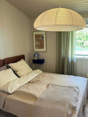 Une chambre à coucher avec un lit, une table de chevet avec une lampe, un luminaire suspendu, un tableau encadré au mur et une fenêtre avec des rideaux.