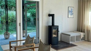 Uma sala de estar moderna com uma lareira a lenha, duas poltronas, um banco e uma janela grande com vista para o verde.
