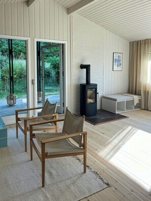Ein modernes Wohnzimmer mit Kaminofen, zwei Sesseln, einer Bank und einem großen Fenster mit Blick ins Grüne.