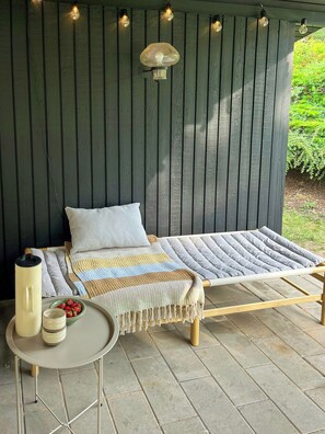 A hammock bed with a striped blanket and pillows, a small table with a candle and a bowl of strawberries, and string lights on a wooden wall.