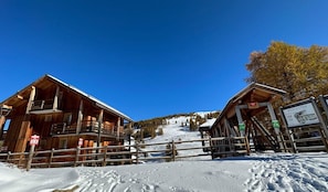 Eine Holzhütte mit Balkon, im Hintergrund eine Seilbahn und eine verschneite Landschaft.