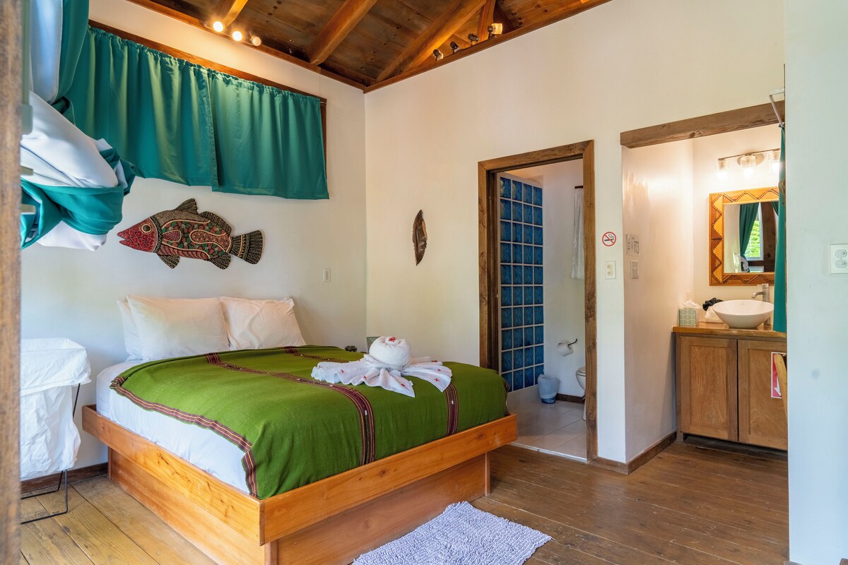 Habitación de hotel con cama de madera, ropa de cama verde, baño blanco con lavamanos y vista al exterior.