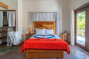 Una cama bien hecha con cabecera de madera, ropa blanca de cama y una manta roja. Hay una pequeña mesa con una lámpara y un cartel encima, y una ventana con cortinas.