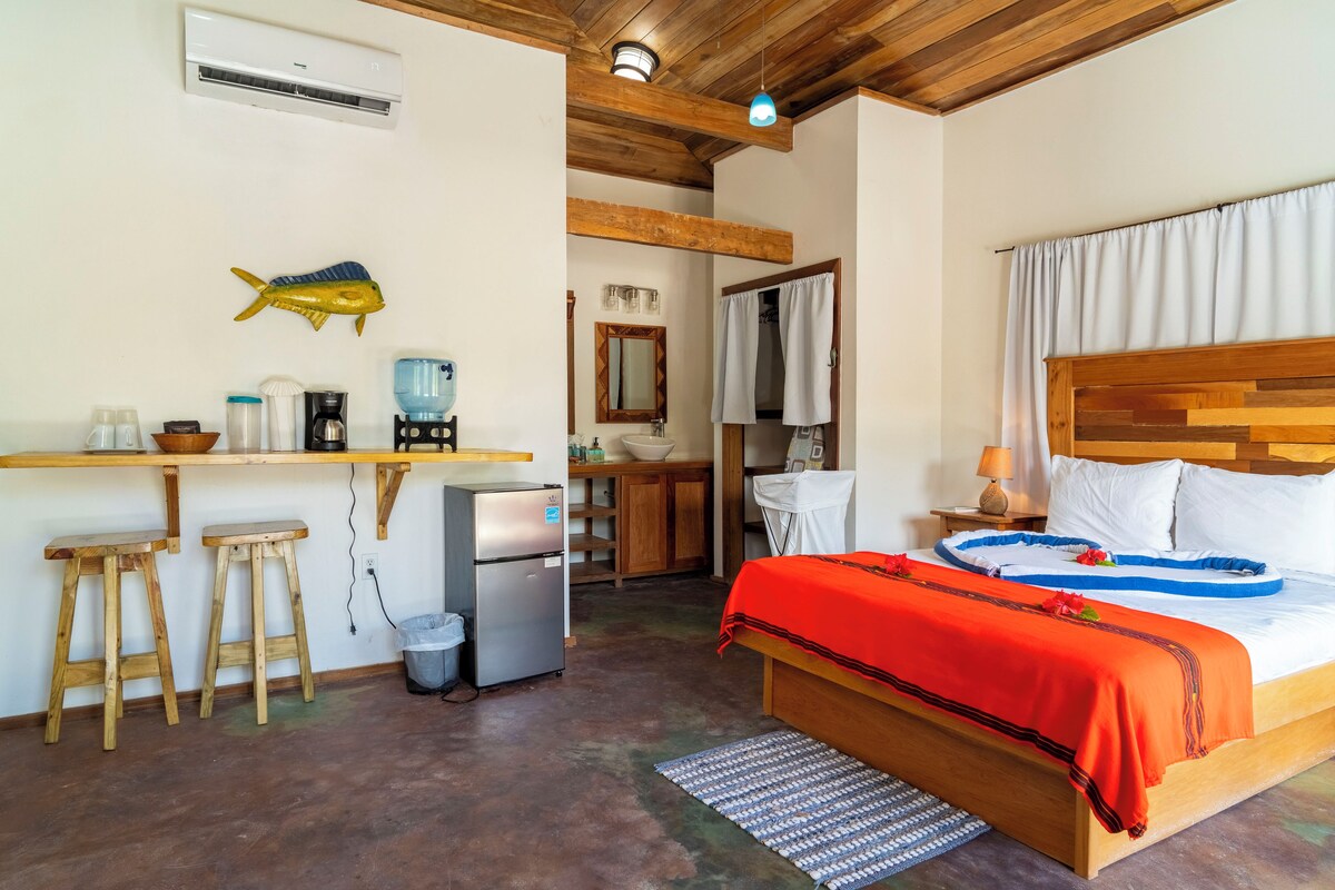 Habitación de hotel pequeña con cama, escritorio, silla, kitchenette y baño.