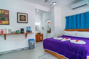 A hotel room with a bed, a shelf with toiletries, a waste bin, and a bathroom visible through an open door.