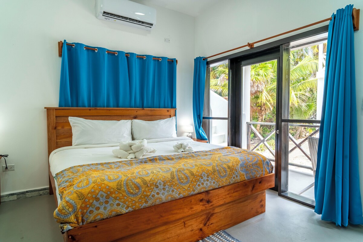 Un dormitorio con cama de madera, ropa de cama blanca, un plaid estampado y vistas a la vegetación a través de un ventanal.