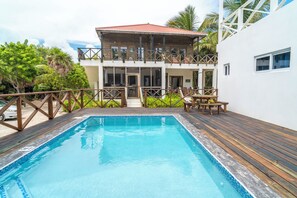 Una zona de piscina con terraza de madera, un edificio de dos pisos con techo rojo y una pared blanca.