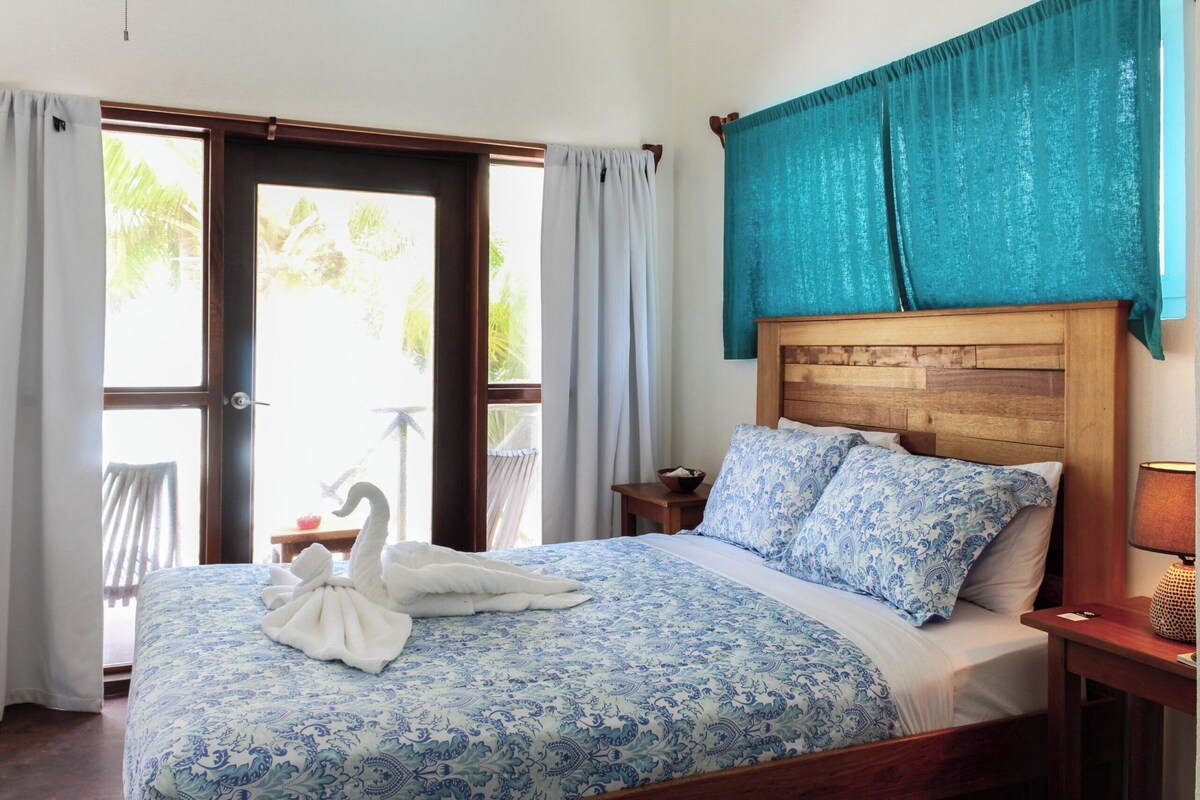Habitación de hotel con cama, cabecera de madera, mesita de noche, lámpara y vista al exterior por una puerta.