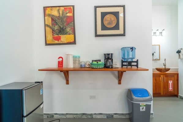 A kitchen area with a wooden shelf holding a coffee maker, a water dispenser, and a trash bin.