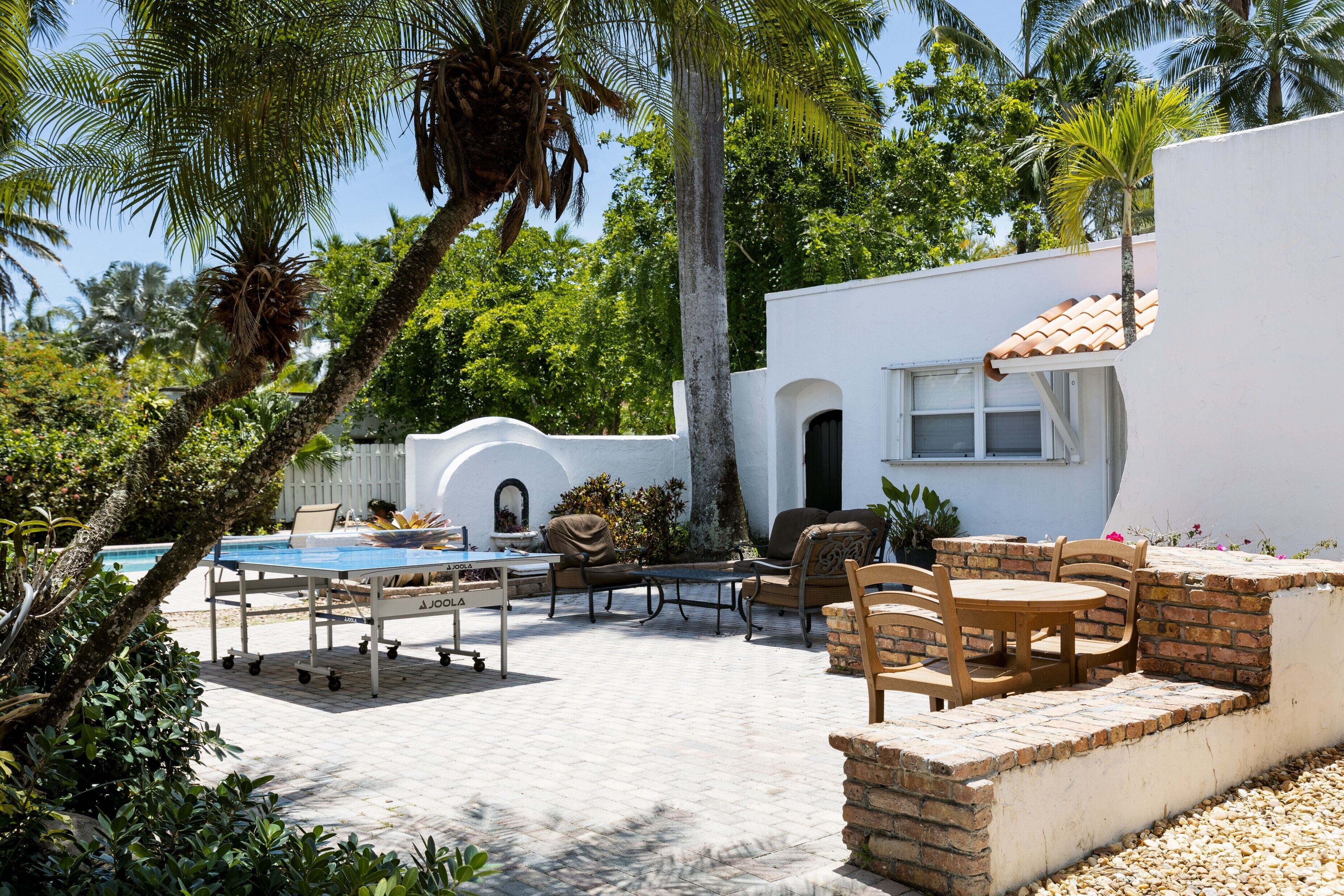 A poolside area with a table and chairs, surrounded by lush greenery and a white building with a tiled roof.