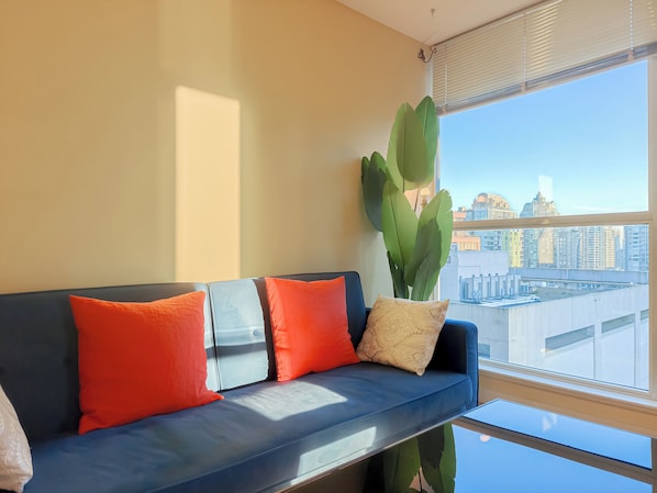 Un salon moderne avec un canapé bleu, des coussins orange à motifs, une grande plante verte et une vue sur la ville par une grande fenêtre.