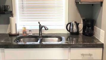 A kitchen with a double sink, white cabinets, a window with blinds, a coffee maker, and a shelf with items.