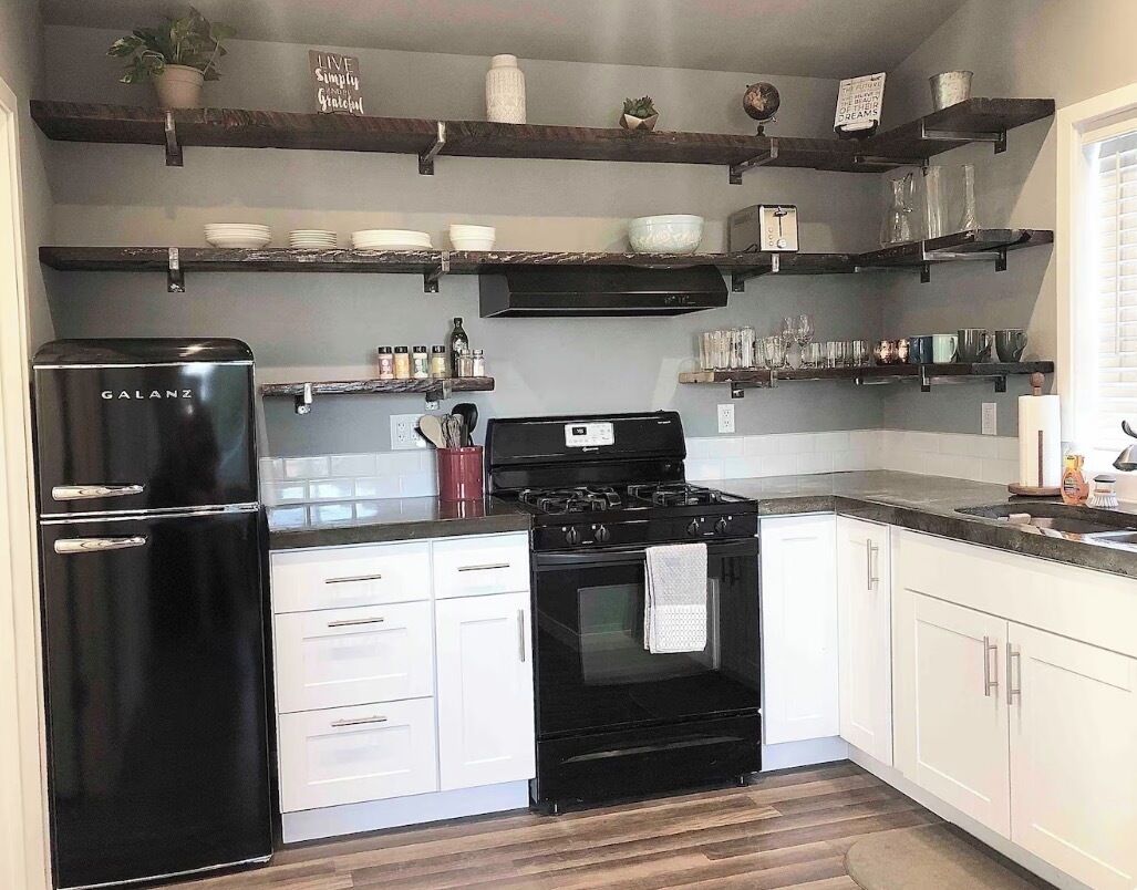 Une cuisine avec des meubles blancs, un réfrigérateur noir, une cuisinière noire et des étagères en bois au-dessus, où sont rangées la vaisselle et les verres.