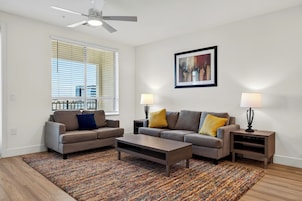 A modern living room with a sofa set, a coffee table, two side tables with lamps, a ceiling fan, and a large window with blinds.