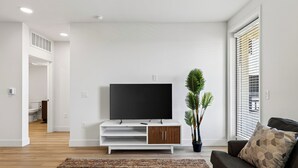 A modern living room with a flat-screen TV on a white stand, a potted plant, a brown sofa, and a patterned rug.