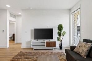 A modern living room with a flat-screen TV on a white stand, a potted plant, a brown sofa, and a patterned rug.