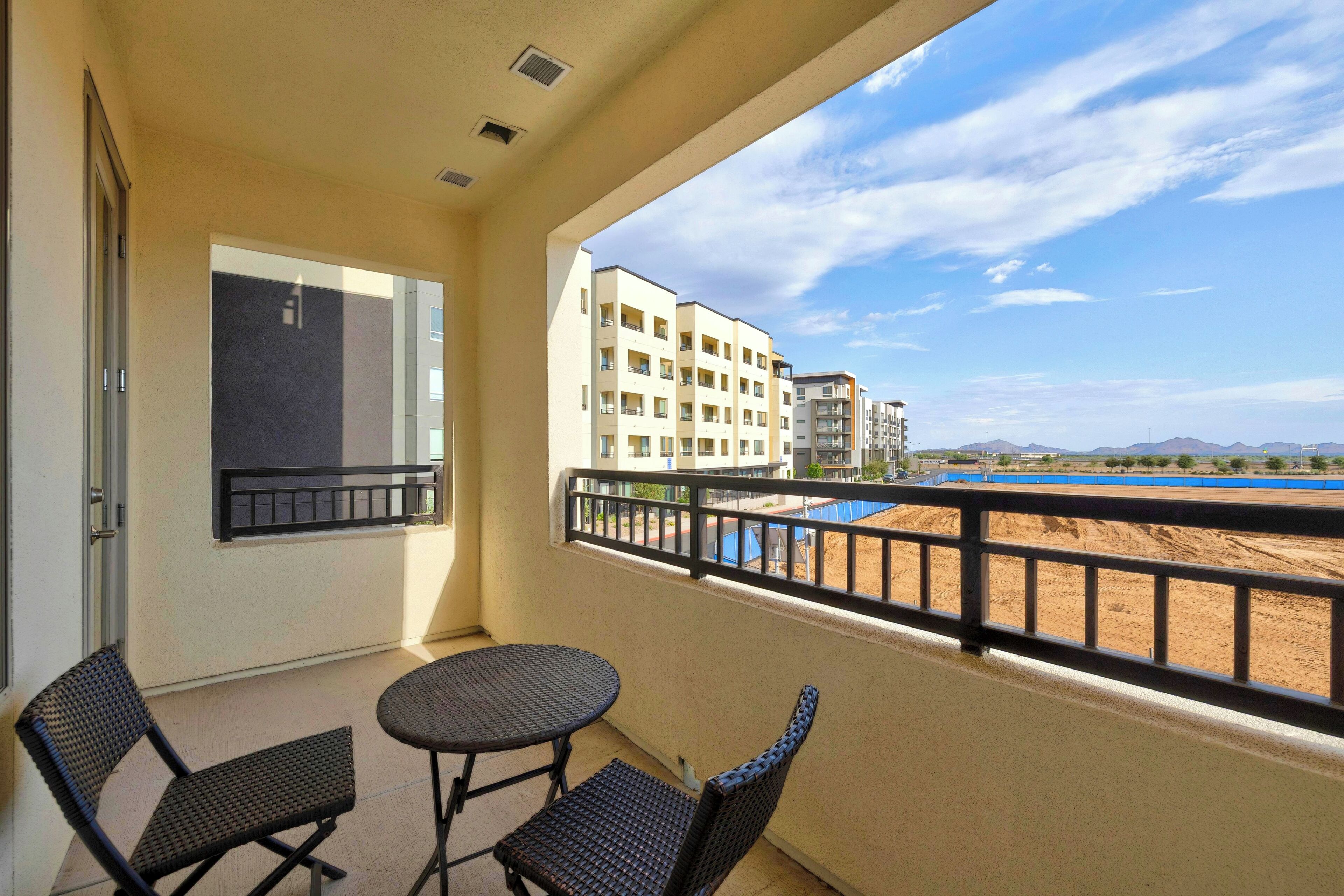 A balcony with two wicker chairs and a small table, overlooking a swimming pool and buildings.