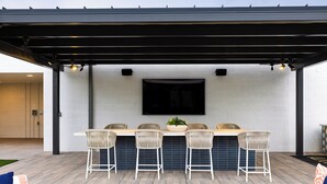 An outdoor patio with a black pergola, a white brick wall, and a flat-screen TV mounted above a bar area with four wicker chairs and a central table.
